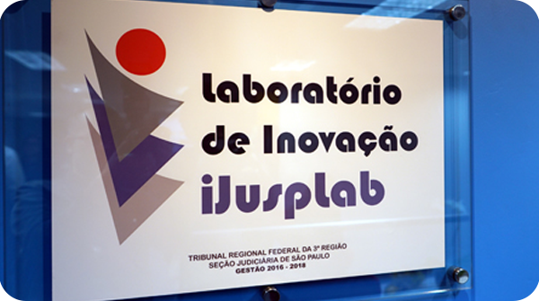 Descrição: Fotografia que mostra uma identificação, na porta, do Laboratório de Inovação iJusplab. No lado esquerdo vemos uma logo e a porta tem cor azul royal. 